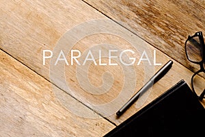 Top view of eyeglasses,notebook and pen on wooden background written with PARALEGAL