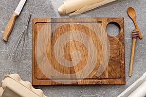 Top view f wooden empty cutting board,knife,whisk,baking paper,eggs paper box,wooden spoon,honey dipper on the grey table