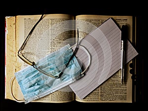 Top view of a facemask with glasses, paper, and a pen on an open old dictionary