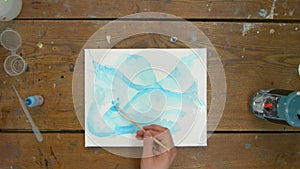 Top view of female artist paints an abstract picture, he uses blue paint on wet canves and distribute it with paintbrush