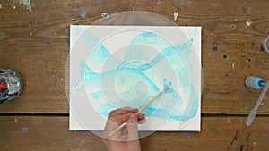 Top view of female artist paints an abstract picture, he uses blue paint on wet canves and distribute it with paintbrush