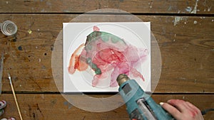 Top view of female artist paints an abstract picture, she uses dryer to distribute paints and dry it