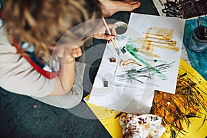 Top view of a female artist sitting on the floor in the art studio and painting on paper with a paint brush. A young woman painter