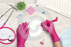 Top view of female doctor hands  in gloves write in a notebook. Medical workplace with stethoscope, keyboard, thermometer and