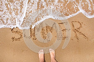 Top view of female feet standing on the beach sand with wave motion coming.Word summer writting on the sand.Summer travel and