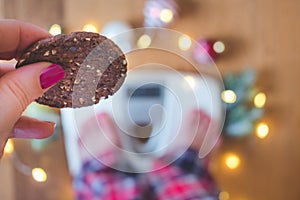 Top view of a female hand holding a cookie and scales with Christmas decorations and lights on wooden background