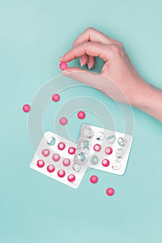 Top view of female hand holding pink prescription pill next to blister packs over pastel blue background.