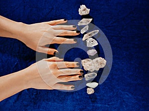 Top view female hands with black nails lying on table with gem stones. Blue velvet for background