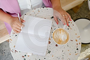 Top view of female hands with cup of coffee sitting at workplace with notebook