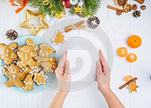 Top view female hands holding empty greeting card under white wooden table with gingerbred cookies, christmas tree branches with d