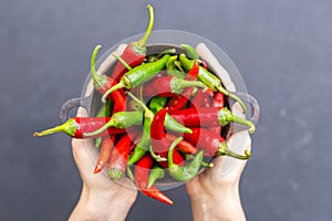 Top view of female hands holding green and red hot chili peppers in a rusty bowl on a dark tabletop