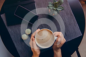 Top view female hands holding hot cup of coffee above workspace with notebook, smartphone, sweets and green potted plant on dark