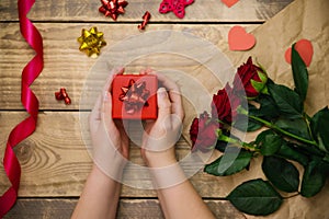 Top view, female hands holding a red gift box. Festive atmosphere, red roses and bows on a wooden background