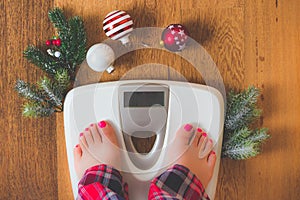 Top view of female legs in pajamas on a white weight scale with Christmas decorations and lights on wooden background