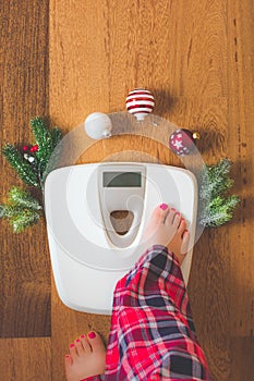 Top view of female legs in pajamas on a white weight scale with Christmas decorations and lights on wooden background