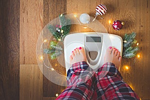 Top view of female legs in pajamas on a white weight scale with Christmas decorations and lights on wooden background photo