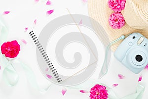 Top view feminine background with flowers and girl`s items. Flat lay blank paper notepad, modern instant camera, staw hat, ribbon