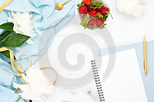 Top view feminine desk workspace with peony flowers, blank paper notepad, bowl of strawberries. Flat lay style composition with