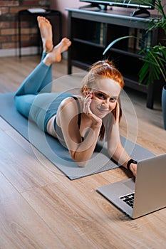 Top view of fit smiling redhead young woman lying on floor and using laptop computer at apartment