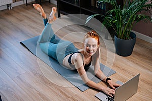 Top view of fit smiling redhead young woman lying on floor and using laptop computer at apartment