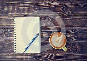 Top view, flat lay latte art coffee in a yellow cup with a blank notebook, and a blue pen on the old wooden table background