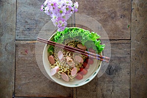 Top View Food Photo of Dried Instant Noodles with Sausages, Cherry Tomatoes, Peanuts and Lettuce on a Wooden Table with Flowers