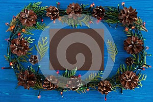 Top view on frame from Christmas lights, fir branches, pine cones and brown sheet of paper on the blue wooden background.