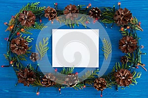 Top view on frame from Christmas lights, fir branches, pine cones and white sheet of paper on the blue wooden background.