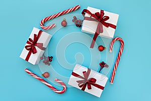 Top view on frame from gift boxes wrapped white paper and red ribbon decorated berries, balls and candy canes on blue background.