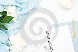 Top view frame of white peony flowers, blank paper notebook, blue scarf and women accessories on marble background. Minimal flat