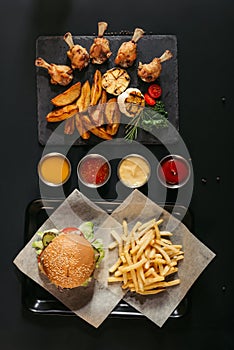 top view of french fries with delicious burger on tray, assorted sauces and slate board with roasted potatoes, grilled vegetables