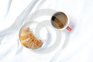 Top view of fresh croissant on white ceramic plate on white tablecloths placed beside hot coffee cup.
