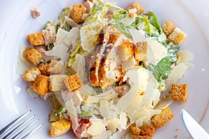 Top view of fresh healthy Caesar salad with grilled chicken breast in a big white plate with fork and knife aside.