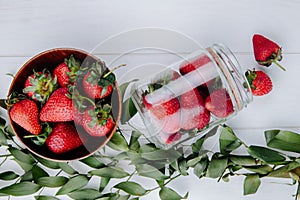 top view of fresh ripe strawberries in a wooden bowl and strawberries scattered from a glass jar and green leaves on white wooden