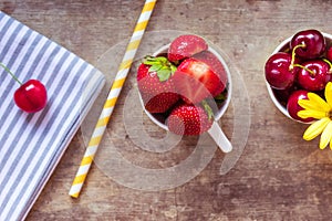 Top view of strawberries, cherries and yellow flowers in bowls with a gray cloth and straw on wooden background