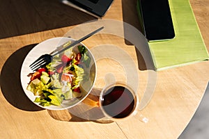 Top view of fresh vegetable salad, laptop, smartphone and glass cup of coffee on wooden table at workplace, nobody