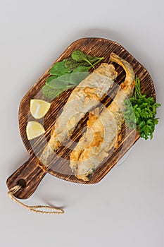 Top view of fried fish navaga served on wooden cutting board with lemon, vegetables and greens on gray background
