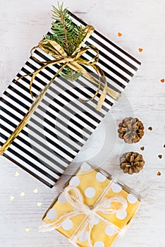 Top view of gift boxes wrapped in black and white striped and golden dotted paper with pine and cones on a white wood background.