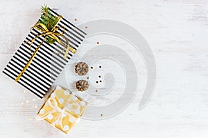 Top view of gift boxes wrapped in black and white striped and golden dotted paper with pine and cones on a white wood background.