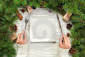 Top view girl holds fork and knife in hand and is ready to eat. Empty white square plate on wooden christmas background. holiday d