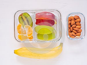 Top view, glass container lunch box of fresh fruit with apple, kiwi, banana, orange and almond on white wooden background. Take aw