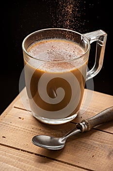 Top view of glass cup of latte coffee with falling cocoa, on wooden table, black background