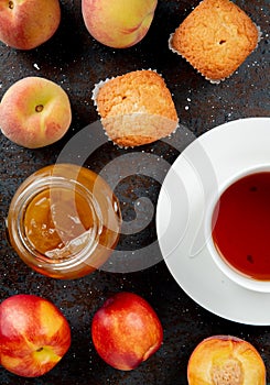 top view of glass jar of peach jam with peaches cupcakes and cup of tea on black and brown background