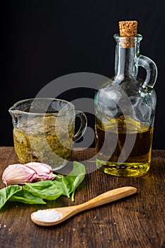 Top view of glass jar with pesto sauce, bottle of olive oil, garlic, basil, wooden spoon with salt, on wooden table, black backgro