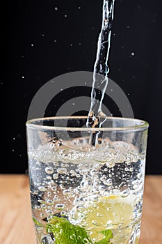 Top view of glass of water with falling water, mint, lemon and bubbles, vertically, on wooden table and black background, vertical