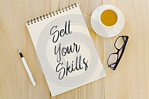 Top view of glasses,a cup of coffee,pen and notebook written with Sell Your Skills on wooden background.Career and job search