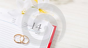 Top view of golden wedding rings and flowers on a calendar, on a white wooden table top.