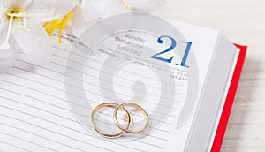 Top view of golden wedding rings and flowers on a calendar, on a white wooden table top.