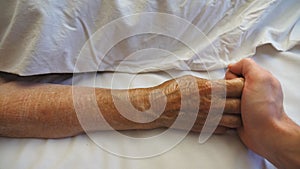 Top view of grandson holds and touches hand of sick mature grandmother in medical clinic. Young man comforts wrinkled