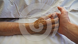 Top view of grandson holds and touches hand of sick mature grandmother in medical clinic. Young man comforts wrinkled